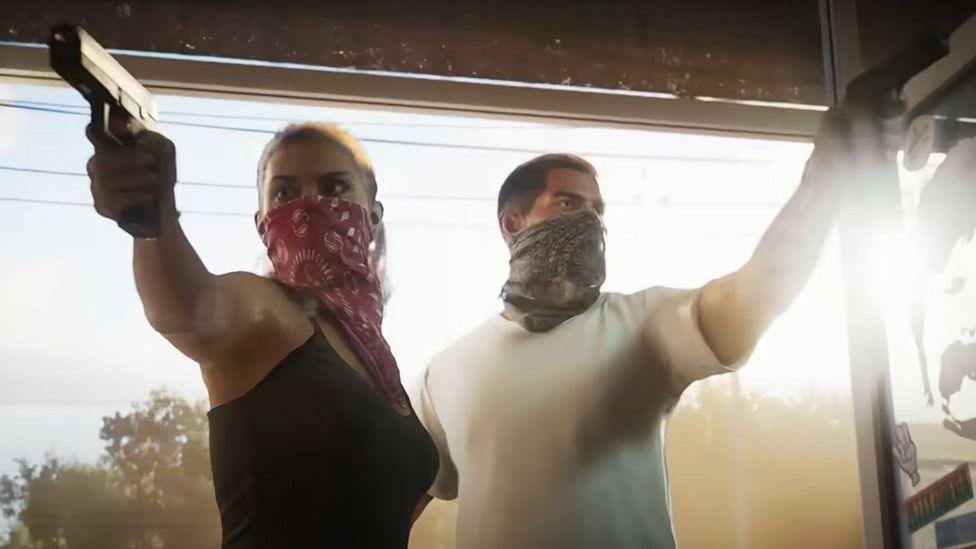 Screenshot shows a man and a woman with their faces covered by Paisley-print bandanas bursting through the glass front doors of a convenience store. Each holds a pistol in their right hand, raised and pointed towards the interior of the shop. the male figure is almost silhouetted by low, bright sunlight coming in from outside. 