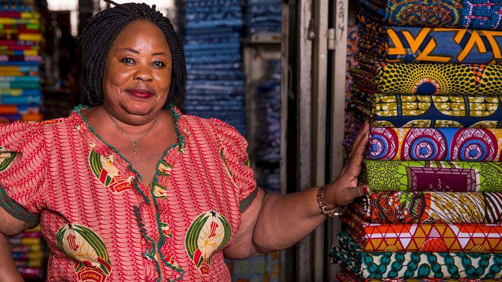 A woman in a red print design shirt standing next to wax print rolls of fabric at a stall in a market in Ghana