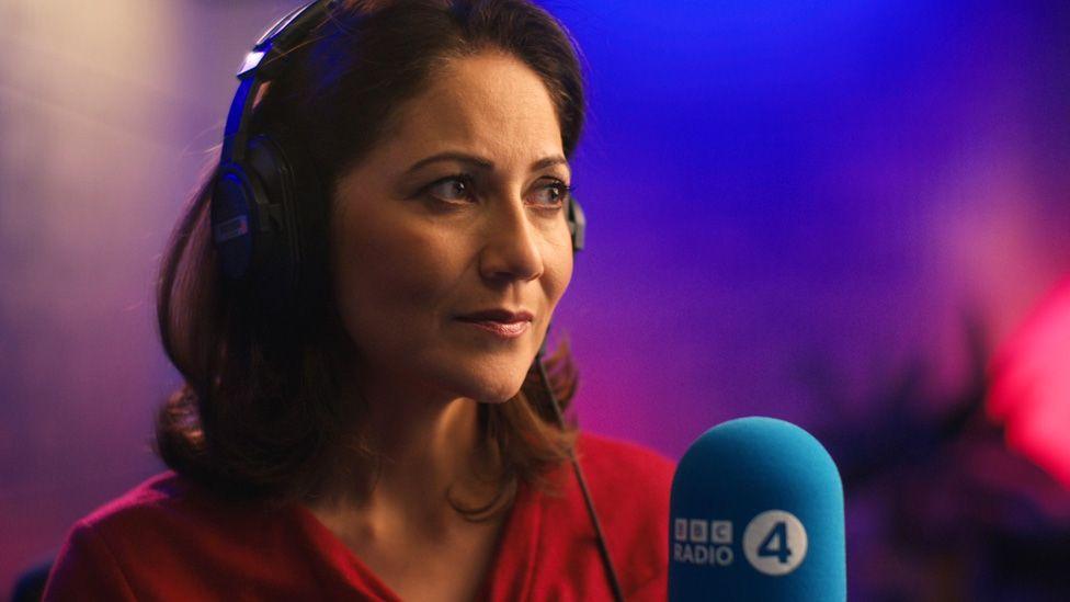 Mishal Husain wearing headphones and in front of a microphone, looking pensively into the distance against a blurred purple and blue background