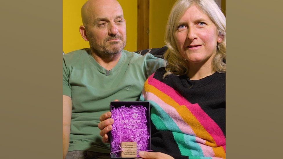Claire Reece and Steve Childerley sitting on a sofa and Claire is holding up a black box with a clear acrylic award in it saying Sense Award and behind it pink shredded paper. Steve is on the left wearing a green T-shirt and Claire on the right with shoulder length blonde hair and wearing a black jumper with a pink, yellow and green stripe across it.