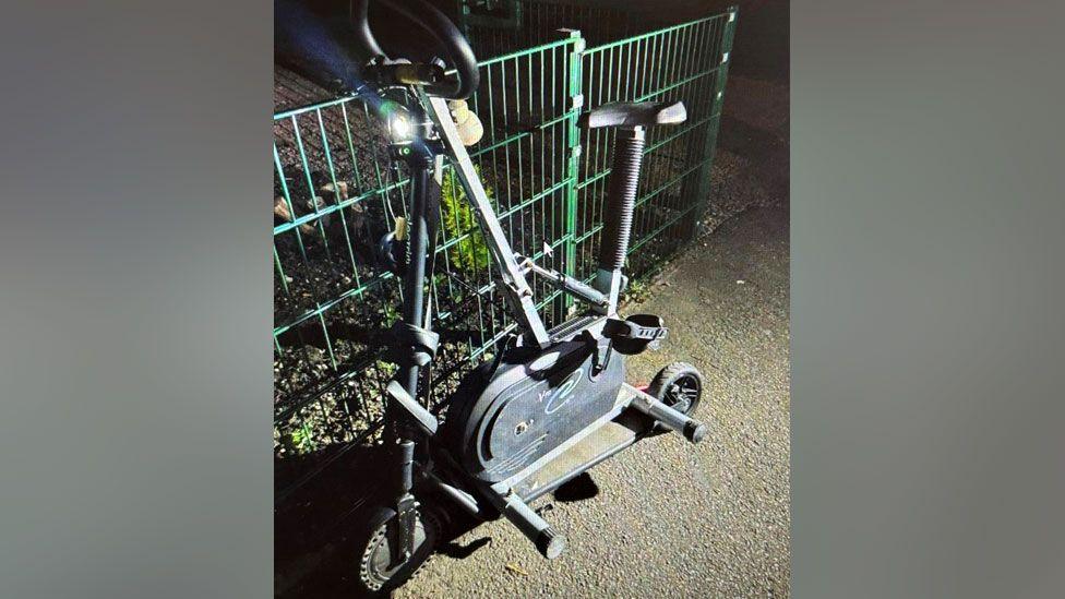 The machine is leaning on a metal fence on a pavement. It has a light on the front.