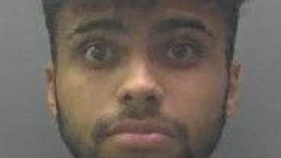 Mugshot of Isaiah Khan. He is brown and has dark hair [ short back and sides and long on top]. He has a dark beard and moustache, which is groomed close to his skin. 