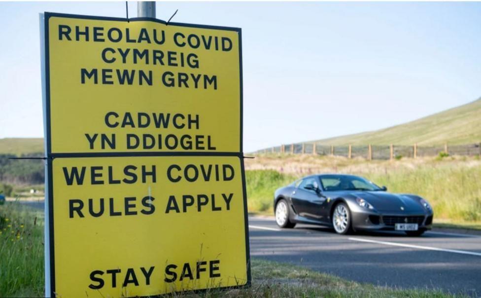 A sign for Welsh Covid rules on the side of the road with a car driving past. 