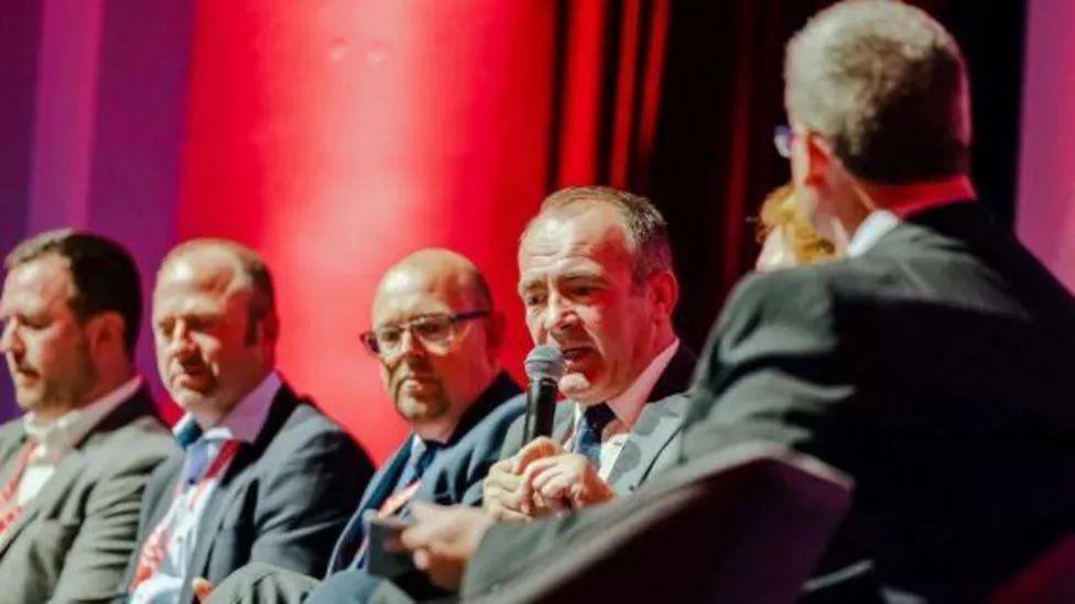 A row of five men in suits, including Chief Minister Alfred Cannan, sitting next to each other one the stage at the 2023 Government Conference. Mr Cannan is speaking into a microphone.