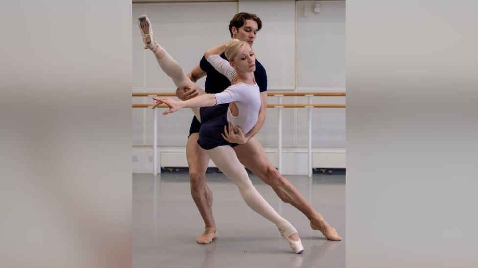 Melissa Hamilton dancing with Lukas Braendsrod in a dance rehearsal studio. Melissa is wearing a white leotard, white tights and white ballet shoes. Her hair is blonde and slicked back Lukas is wearing black t-shirt and shorts. He has brown hair. Melissa's right leg is pointed down leaning forward and her left leg is raised behind her. Gareth has his left arm around her waist supporting her and his right arm is supporting her leg.