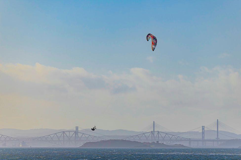 Kitesurfer mid-air with bright kite high above water, with bridges in the distance, and blue sky and clouds.