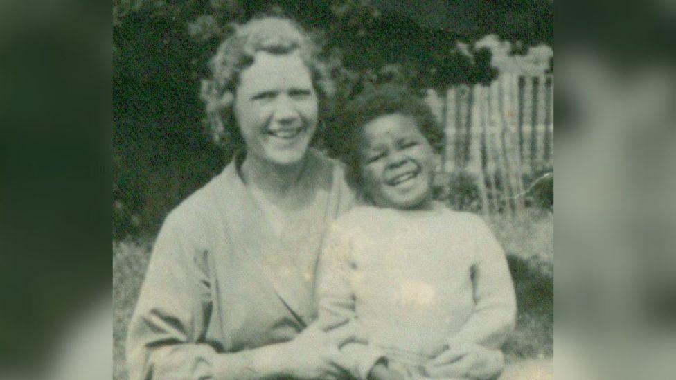 A black and white photo of Dr Stephenson as a young child, wearing a light coloured jumper. He is being held by his foster mother in a garden. Both are laughing and smiling at the camera. 