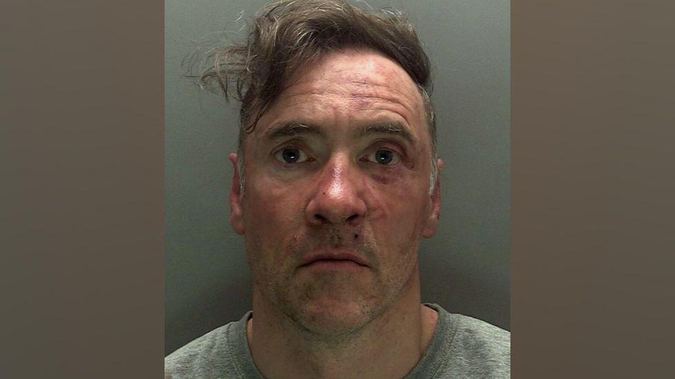 A police custody photo of Paul Doyle wearing a grey jumper and looking stunned. His hair, a long brown fringe, is dishevelled and to the side. His face has red marks on it