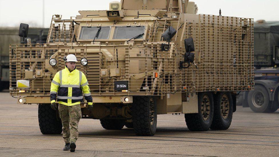 A brown coloured Mastiff armoured vehicle is being driven with a man in high-vis walking in front of it to guide it to a ship.