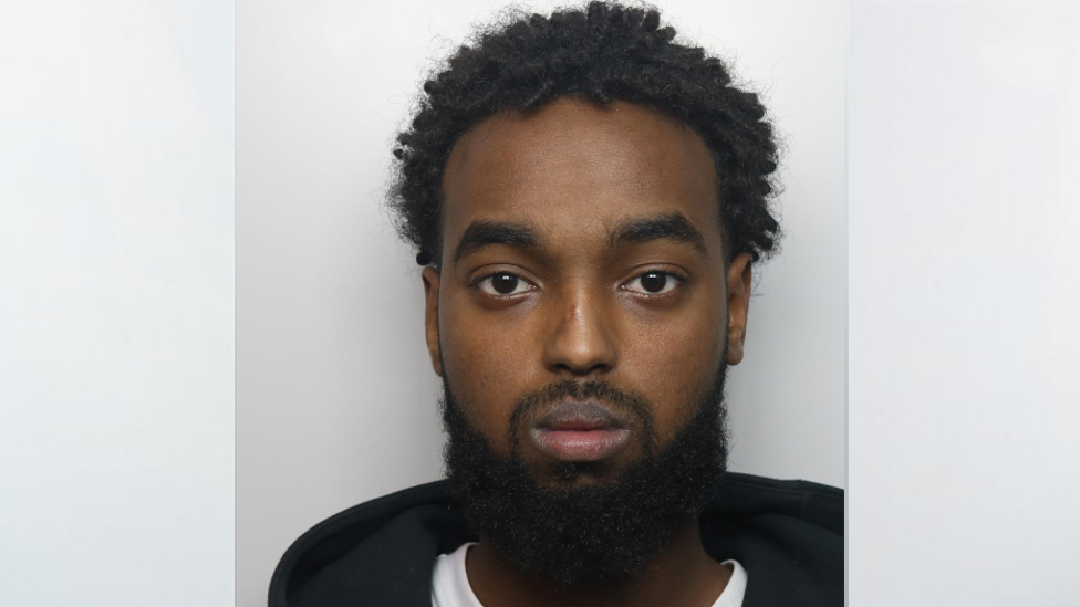 A police mugshot of Mohamed Abdulle, who is staring ahead. He has a short moustache and longer black beard and short, curly hair on top of his head. He is showing no emotion.