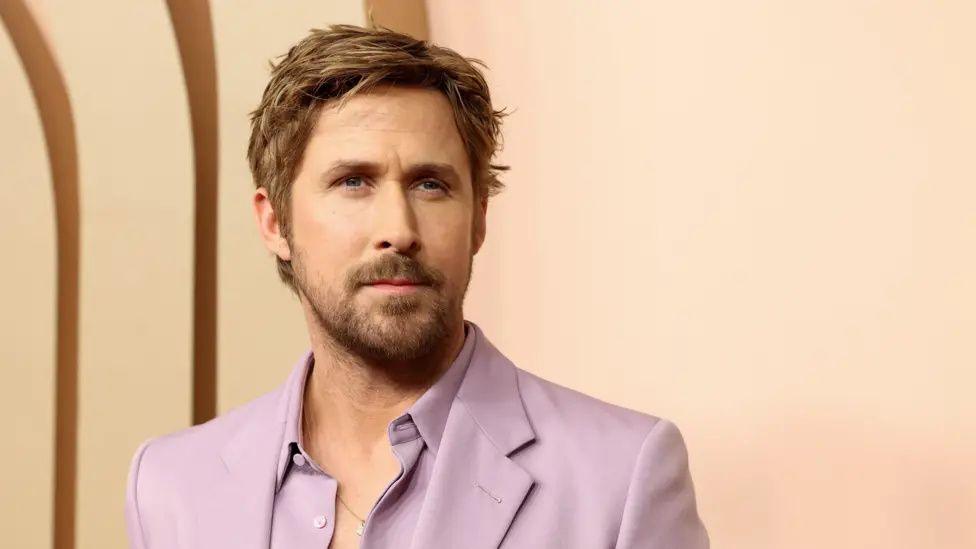 Gosling wearing a lilac-coloured suit and shirt at an official event. The top buttons of the shirt are undone, there is no tie. He is very handsome.