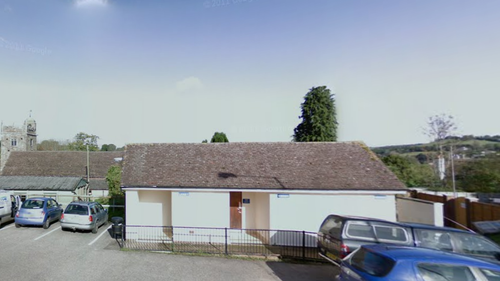 Google street view image of the public toilets in Newton Poppleford. The one-storey block is painted a white colour. Parked cars are to the front and side of the toilet block. 