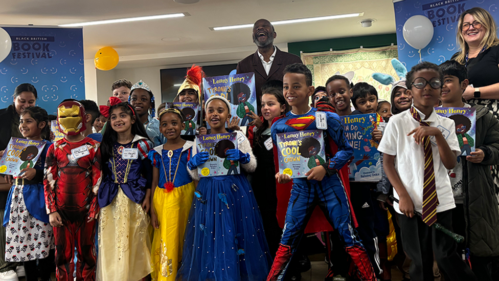 Just over 20 children dressed up in various World Book Day outfits, many of them holding books. They are posing with Sir Lenny Henry in front of banners for the Black British Book Festival