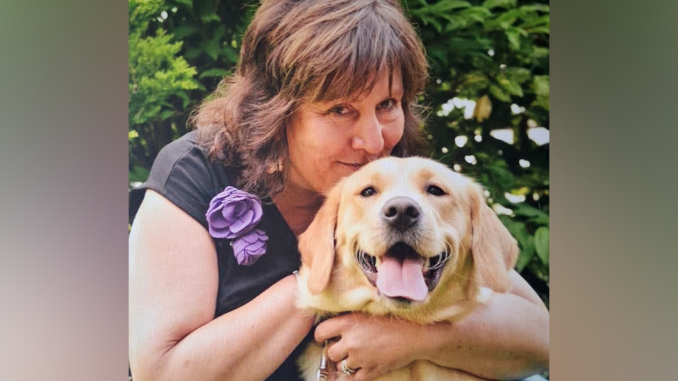 A woman with shoulder length brown hair is hugging a golden retriever guide dog that has its tongue out.