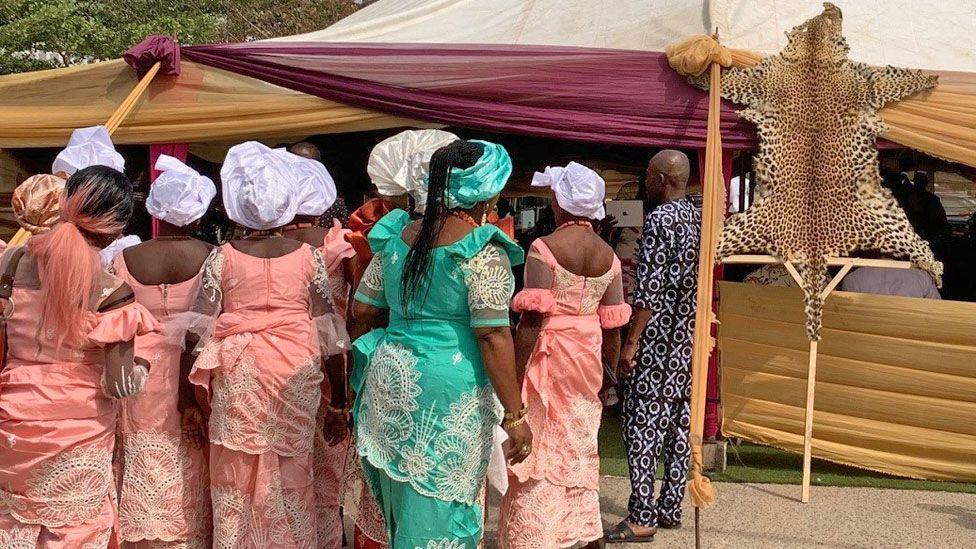 The group of women seen from behind similarly dressed in traditional costumes and headdresses. They are standing in front of a tent on which is propped a fake leopard skin stretched over a pole