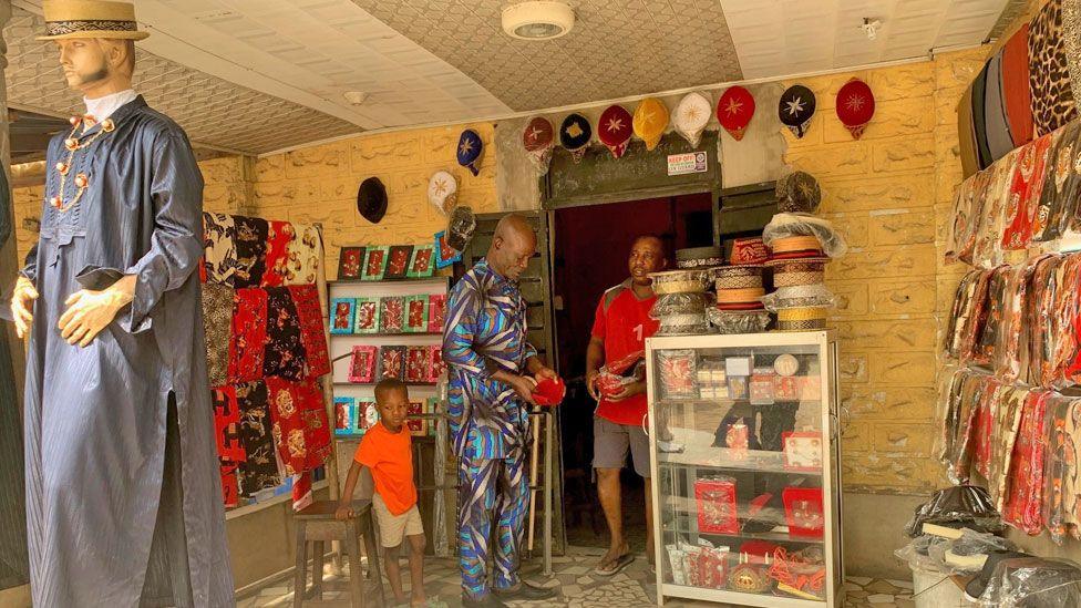 A male customer holding a red cap in a men's clothing shop in Oguta where traditional attire can be seen on display