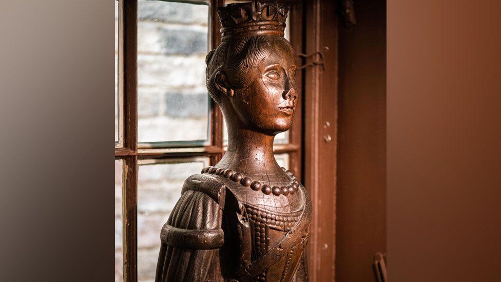 A wooden carving of Queen Victoria, showing her as a young woman. The carving is detailed and reveals a small crown perched on top of her head, her hair pulled back in a bun, beads around her neck and a low-cut dress. The figure is side on and has its back to a window