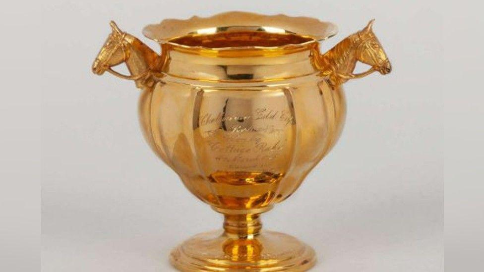 A solid gold cup trophy from the 1940s. The cup is quite bulbous, and the handles are two horse's heads, wearing bridles. The rim of the trophy is fluted.