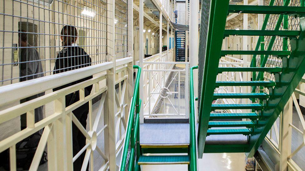 The stairs and corridor of HMP Portland in Dorset are pictured in 2014, with an unidentifiable police officer escorting a prisoner into their cell to the left of frame.