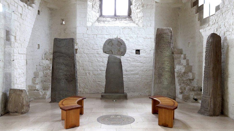 The Celtic stones at St Illtud's Church date from the 8th to 10th century AD. They are large grey stones standing upright in the church, with windows in the background and modern benches in the foreground
