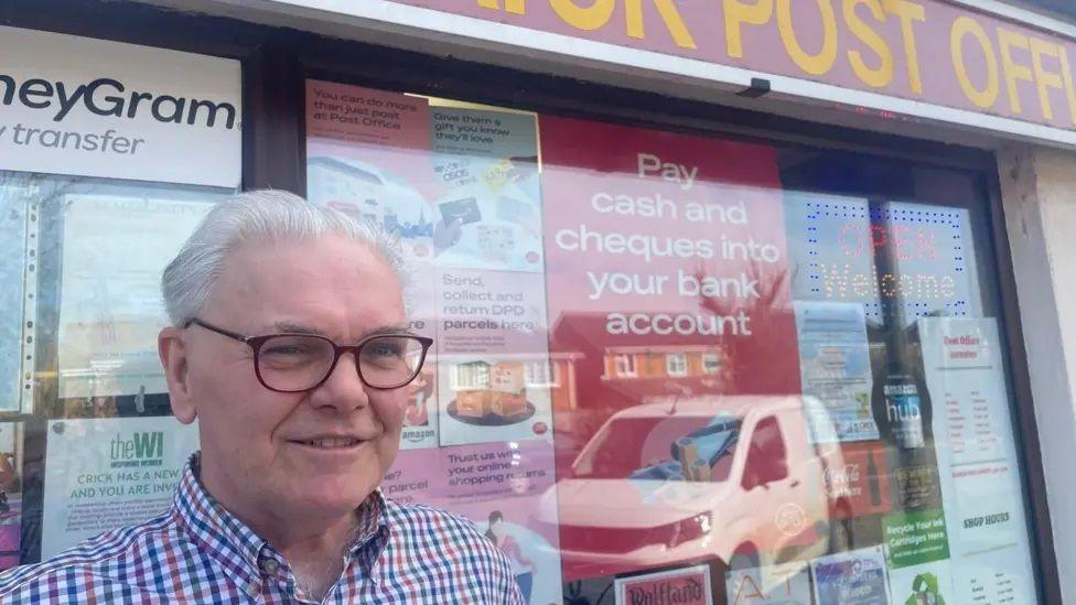 Chris Attridge with short white hair and glasses looking at the camera. His post office is behind him, with several posters in the window and a poster saying "pay cash and cheques into your bank account". The reflection of a white van can be seen in the window.
