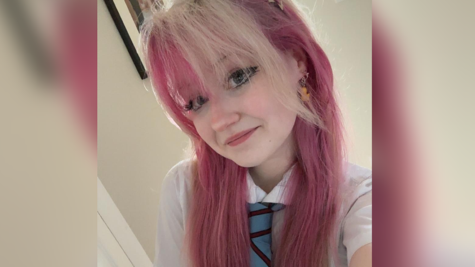 A selfie photograph of Holly with died pink hair wearing a white shirt and a blue and burgundy striped school tie. She has dark eye make up, rubber duck drop earrings and is smiling at the camera.