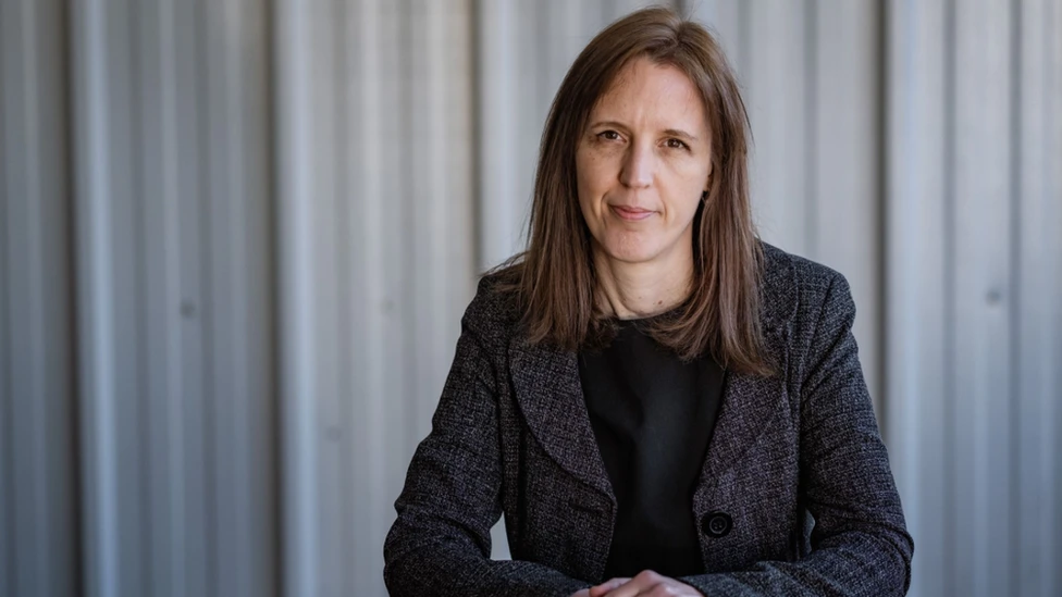 Image of the Green Party leader of Stroud District Council, Catherine Braun, looking at the camera wearing a black top and jacket in front of a grey corrugated metal wall 