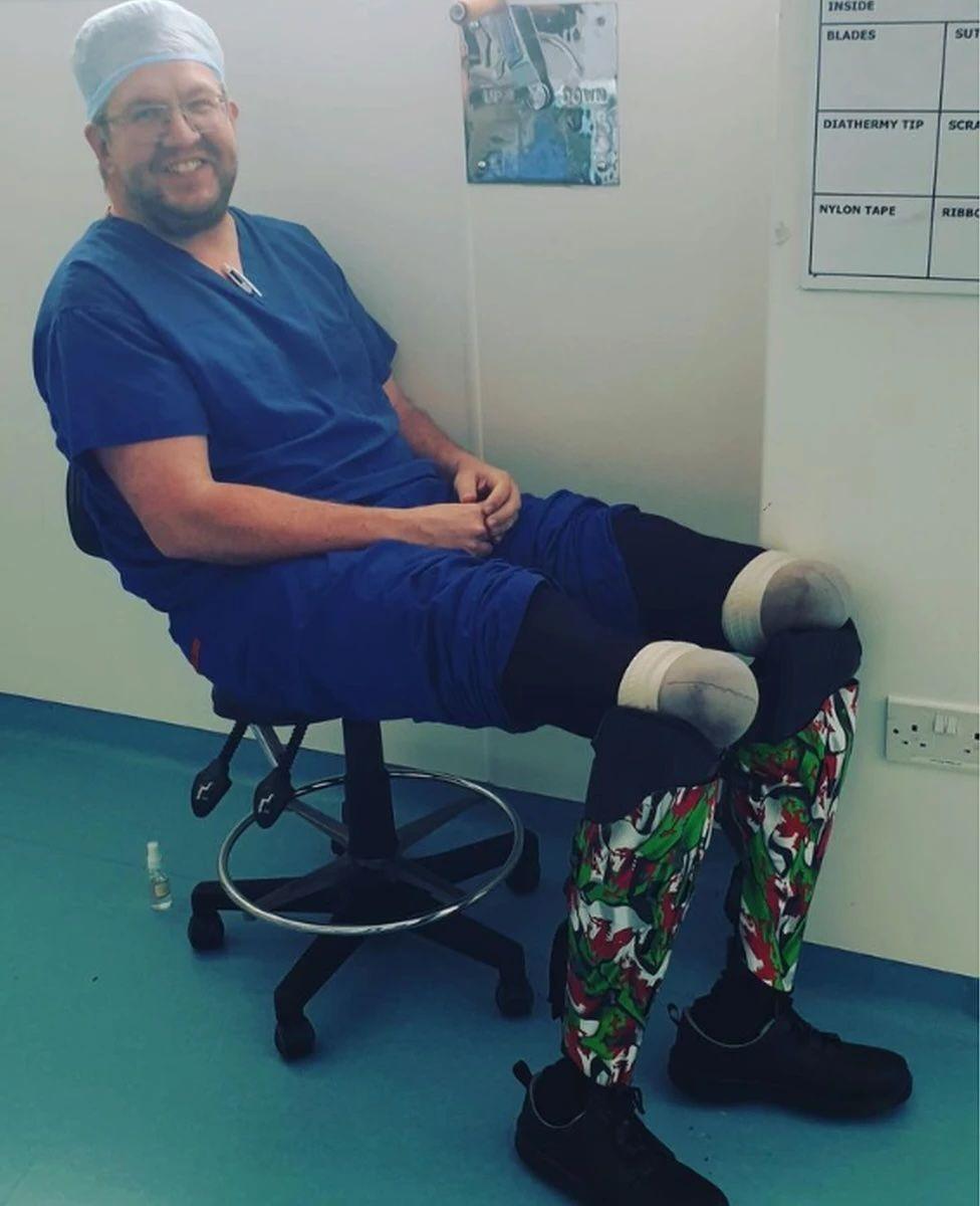 A person with two prosthetic limbs, sitting down on a swivel chair and wearing a blue outfit and cap.