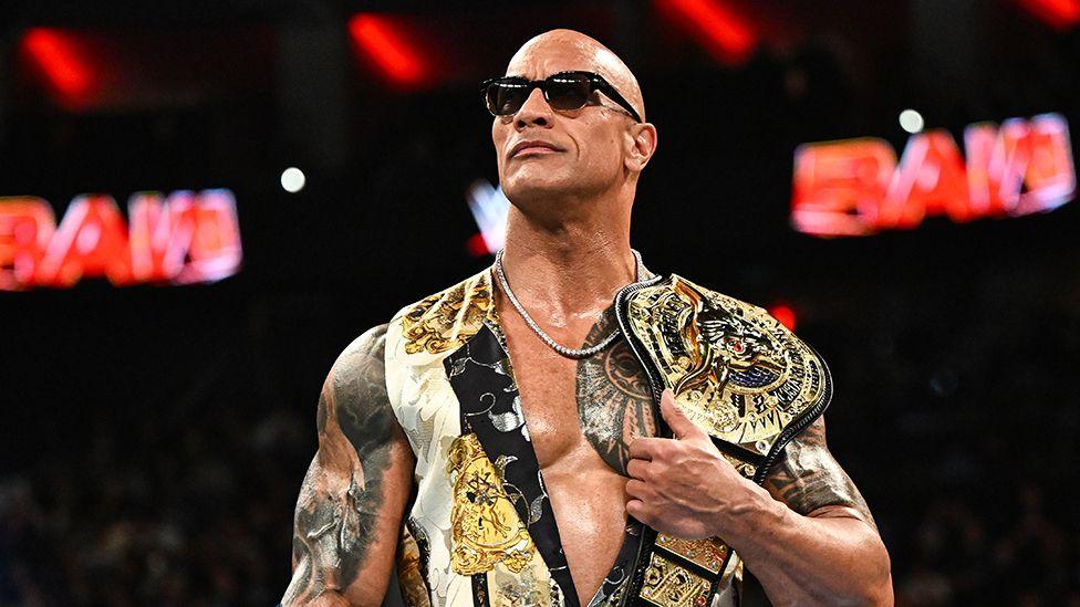 The Rock standing in the middle of the ring, holding a golden title belt over his left shoulder, while wearing black sunglasses, a necklace chain and a white and gold top. Behind him is the "RAW" sign in red.