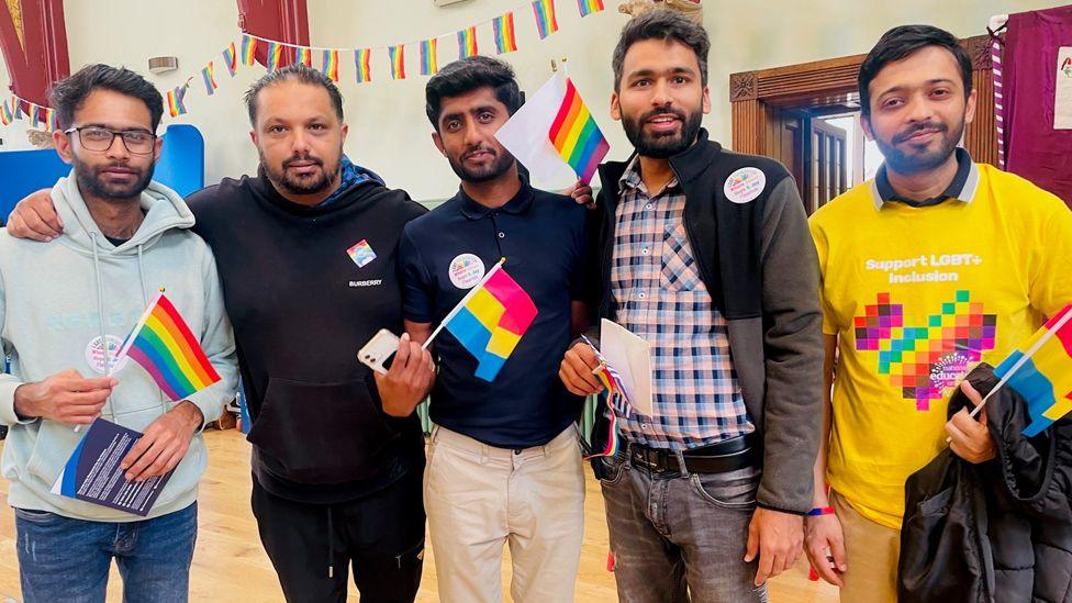 Five men dressed in casual clothing and waving small rainbow flags, smile and look at the camera