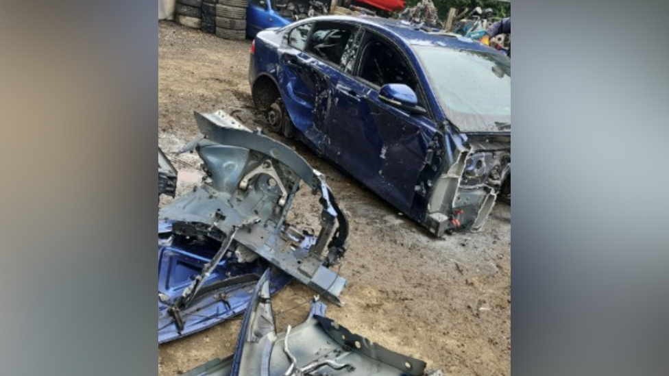 A broken up blue car in a scrap yard with its front missing and parts of its bodywork strewn around it