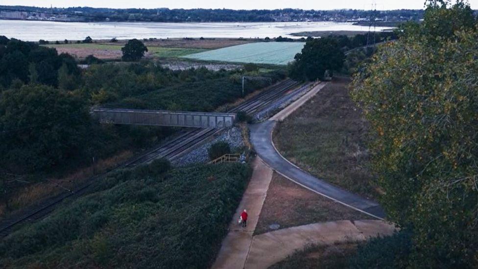 An aerial image of a woman wearing a pink jacket walking a dog. A railway track and expanses of water can be seen, as well as wooded areas.