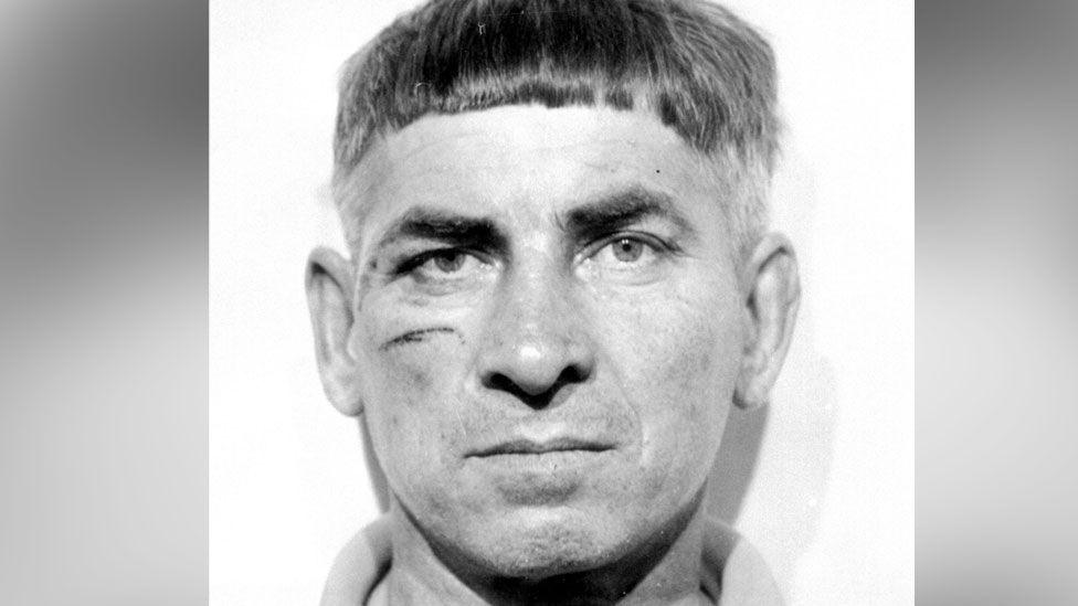 A black and white police mugshot of Peter Cook from 1975 showing a scar on his right cheek. He has short hair and is expressionless.