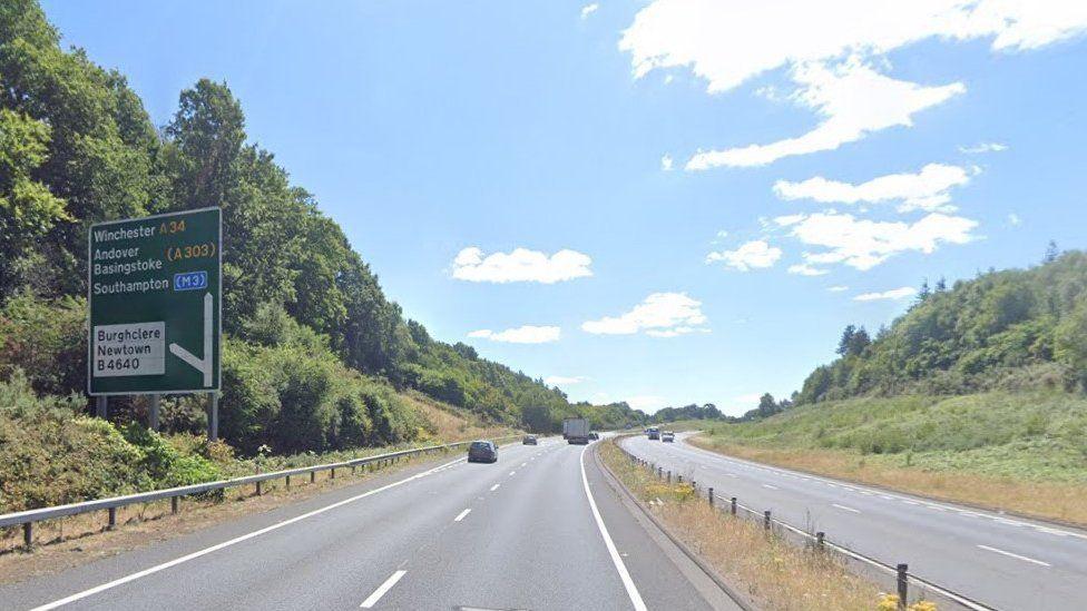 Both sides of dual carriageway with tree-lines banks and large road sign on left