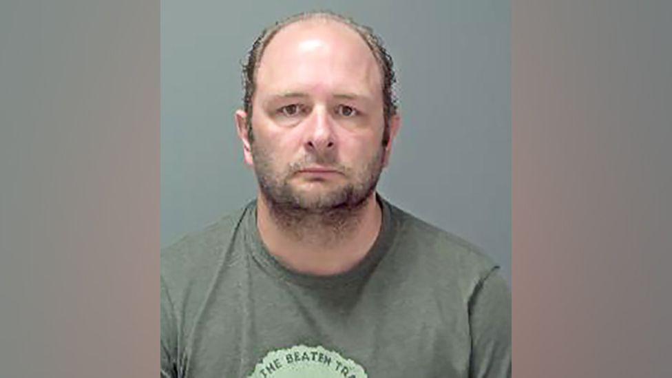 A mugshot of a balding man with facial hair wearing a khaki t shirt starting into the camera