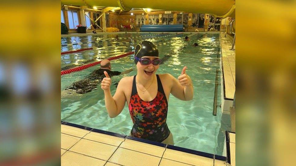 Journalist Eleanor Maslin in a speedo costume with stars on putting both her thumbs up at the end of a pool whilst wearing her goggles and Pudsey swim hat. Lanes and other swimmers can be seen behind her.