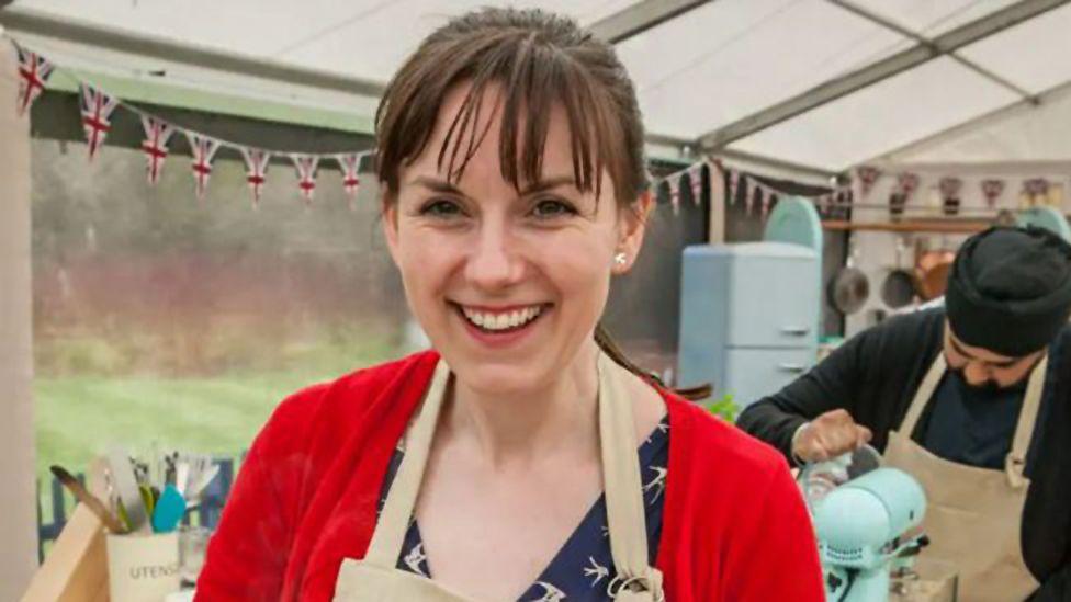 Kate Barmby on the Great British Bake Off set, smiling at the camera. She has brunette hair with a fringe and ponytail. She is wearing a blue top and a red cardigan, covered by a beige apron. In the background are kitchen utensils, a fridge, bunting and another contestant.