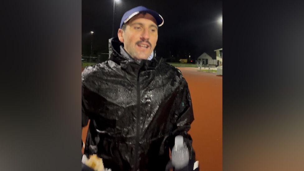 Oliver Gooch running round the track in the middle of the night. He is wearing a blue baseball hat and black coat that is wet through with rain