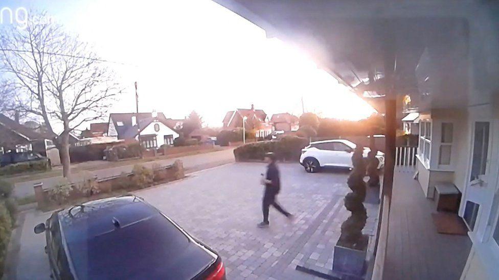 A CCTV still of Luke D'Wit, wearing all black and a cap, walking along the Baxters' paved driveway. There is a black car and a white car parked on the large driveway, which has various shrubs and flowers.