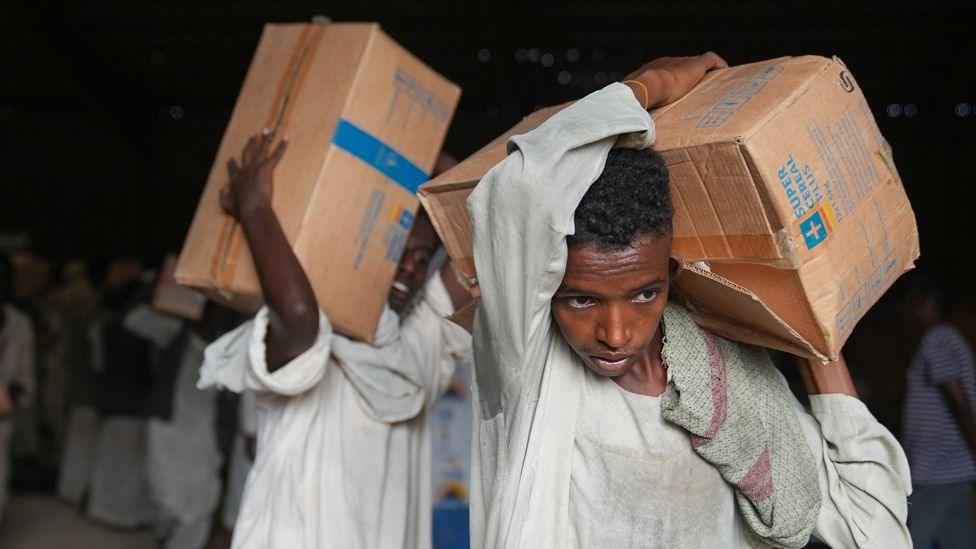 Two men in white clothes carry boxes of aid on their shoulders. 