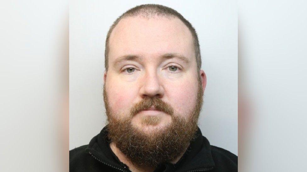A police photo shows a man with a large beard and wearing a dark fleece
