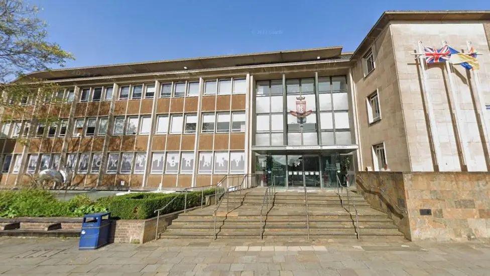 Landscape view of Warwickshire County Council headquarters. It is glass fronted and has stairs leading to the main entrance.