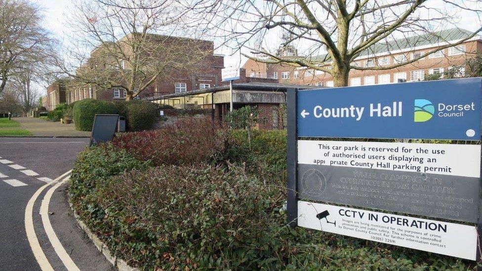 County Hall in Dorchester. In the foreground is a signpost to the large redbrick office building which is surrounded by shrubs.