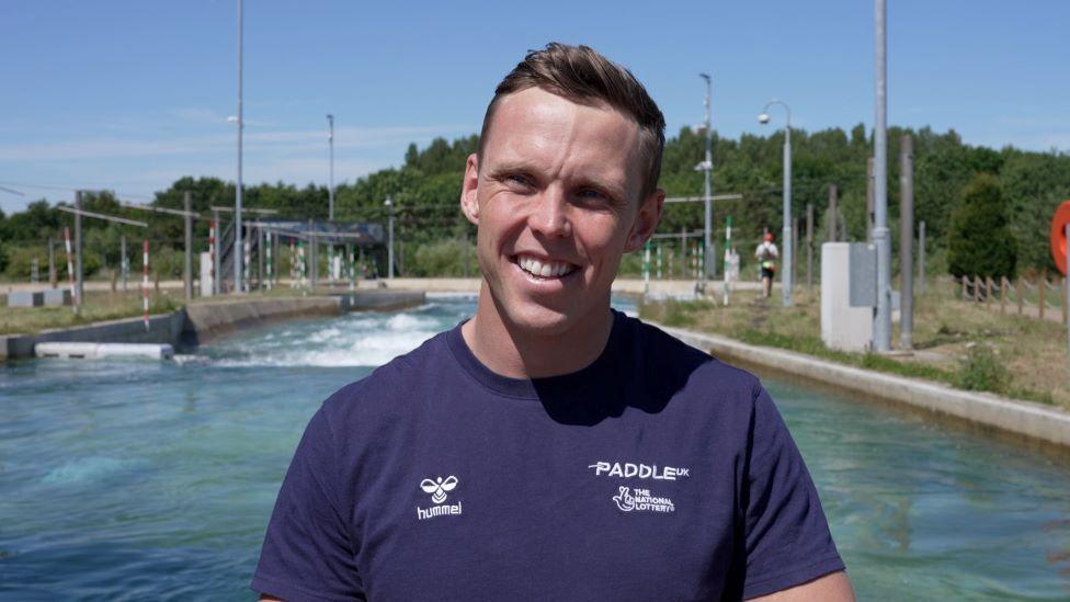 A smiling Joe Clarke wearing a blue T-Shirt looks directly towards the camera, squinting slightly from the sunshine, and standing in front of a slalom course at Lee Valley Water Centre