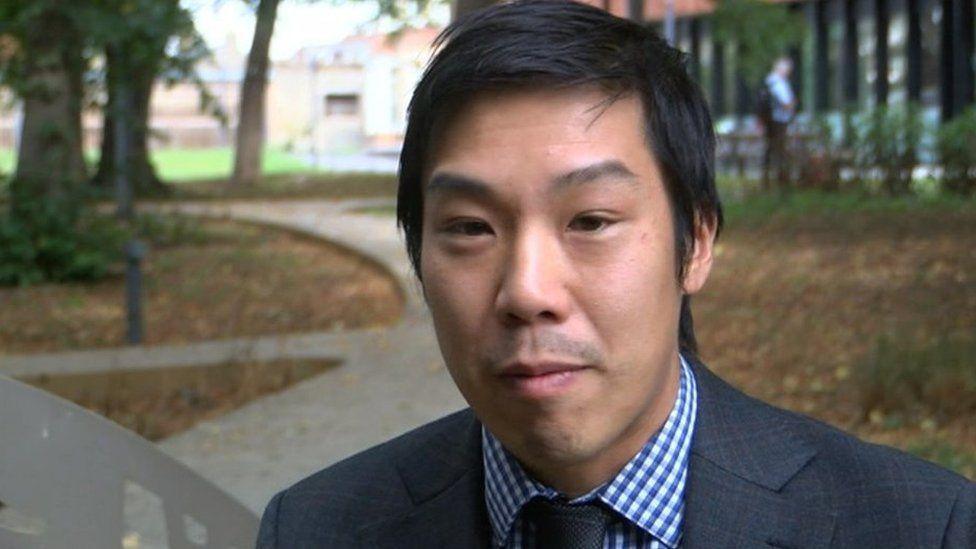 Dr Lennard Lee has medium length black hair, and is wearing a grey blazer, blue tie and a blue and white chequered shirt.