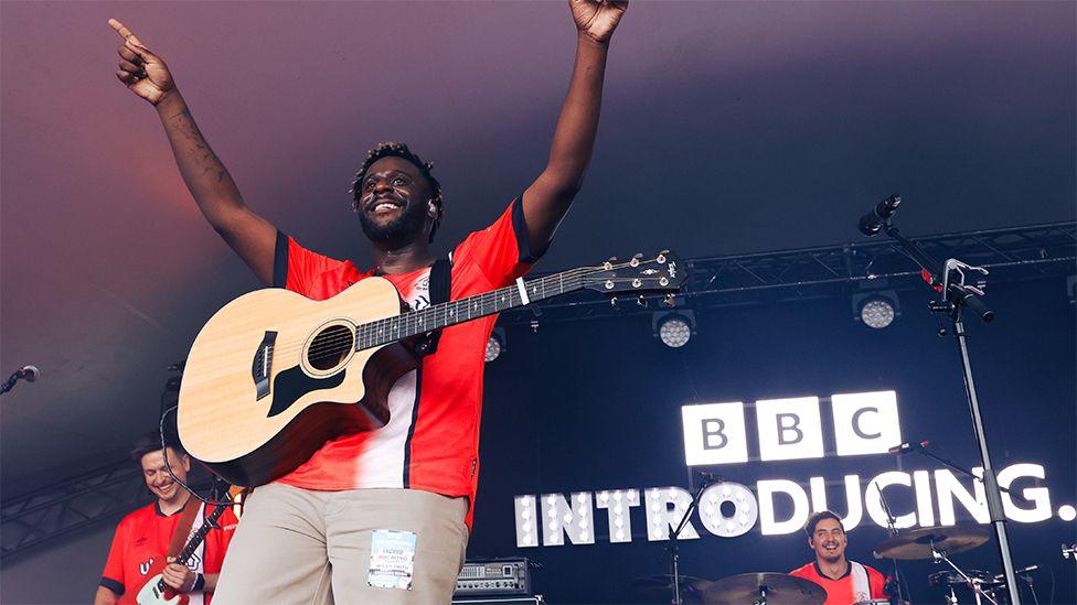 Myles Smith performing on the BBC Introducing Stage at Radio 1's Big Weekend in Luton. Myles is wearing a red Luton football jersey with a white stripe, and brown trousers. He has a guitar around his neck and both arms pointed up in the sky. Behind him are two members of the band and "BBC Introducing" lit up in white font and a black background.