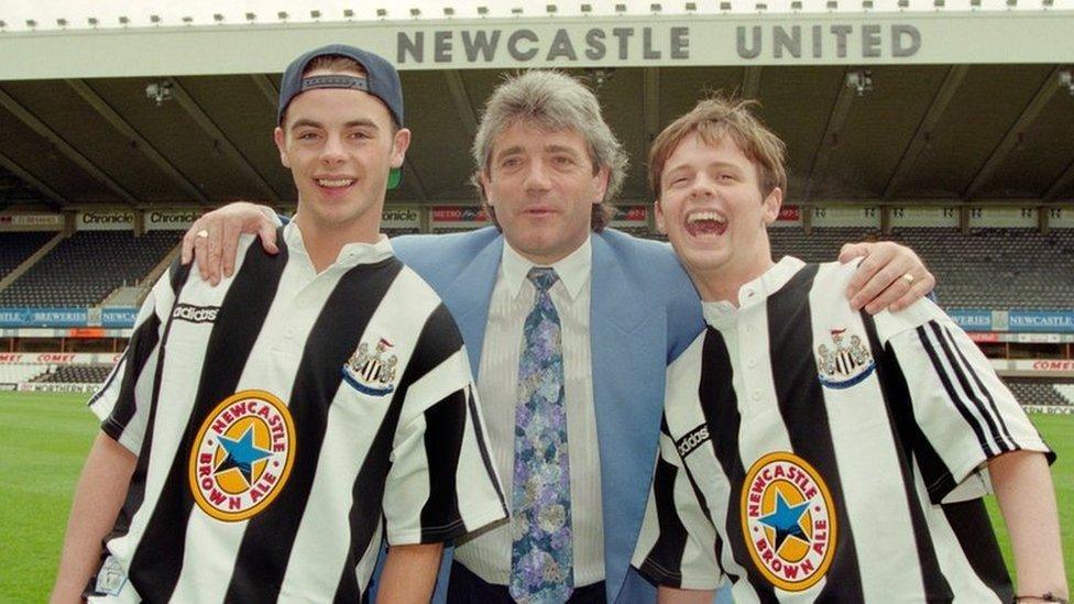 Kevin Keegan posing with TV personalities Ant and Dec at St James' Park in the 1990s. Ant and Dec are both wearing black-and-white striped shirts with a Newcastle Brown Ale logo on the front. It's a yellow circle with a blue star.