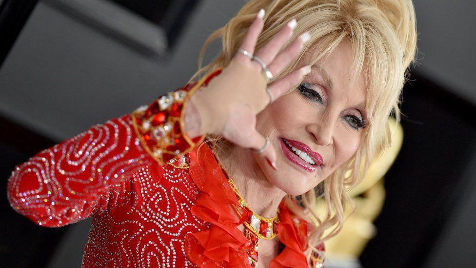 Dolly Parton, with blonde hair and wearing a red dress encrusted with shiny studs, waves at the camera.