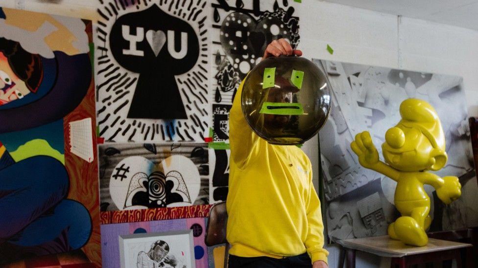 Bristol artist Sickboy is photographed in his studio in front of a wall of paintings he has done. He is wearing a yellow jumper and holding up a goldfish bowl to cover his face.