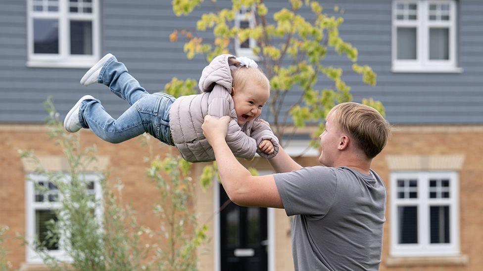 Alex Tarling, who's wearing a grey T-shirt and has short, blond hair, is swinging his small daughter around in the air. She's wearing jeans and a puffy coat and is smiling.